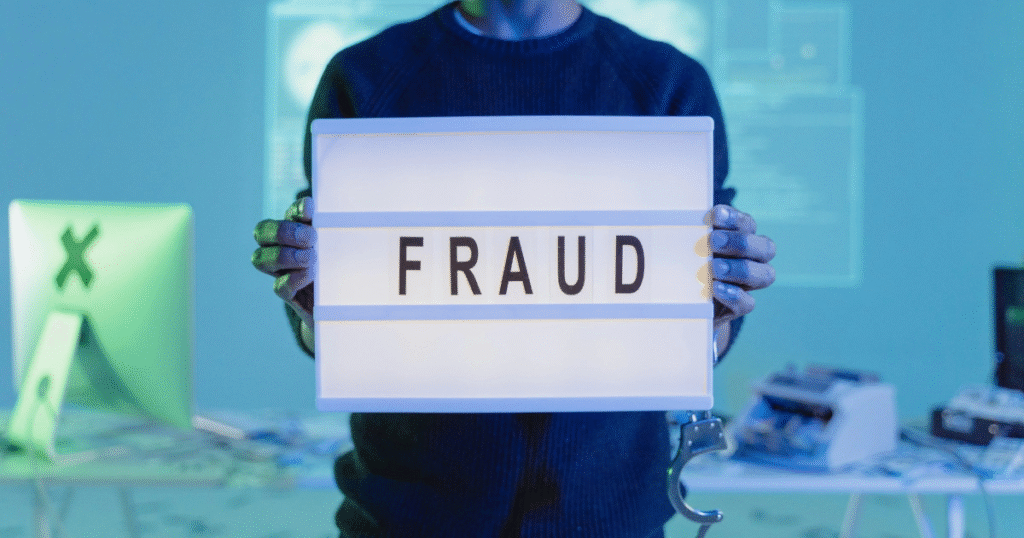 AI in cybercrime illustrated by a person in a dark sweater holds up a glowing lightbox that reads "FRAUD," with a pair of handcuffs dangling from their wrist and computer monitors in the background.